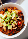 A white bowl of butternut squash chili topped with tortilla strips, avocado, and red pepper flakes