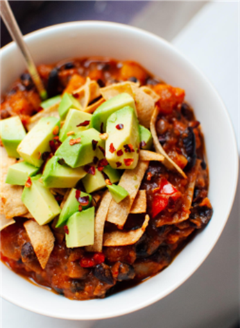 A white bowl of butternut squash chili topped with tortilla strips, avocado, and red pepper flakes